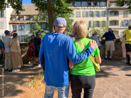 Rear view of a mature couple. The man has his arm around his wife. They are on a group walking tour and stop to take in the views of La Petite, France. Both are dressed casually.