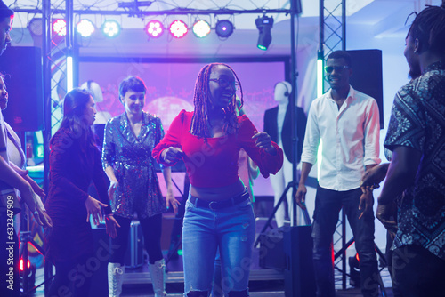 Woman clubber showing moves at improvised dance battle for diverse friends in nightclub. African american dancer partying on dancefloor illuminated with stage spotlights in club