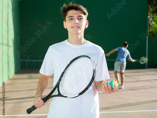 Smiling young hispanic guy in sportswear with racket and colorful rubber frontenis balls in hands posing on outdoor fronton court, ready for friendly match on summer day