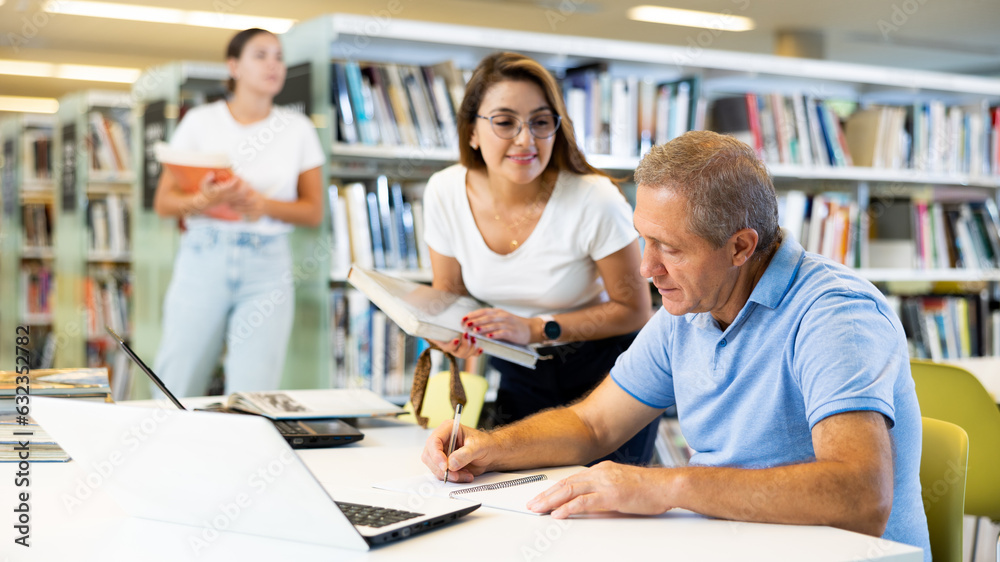 Obraz premium Concentrated mature man spending time in library, reading books and making laptop. Self-education concept