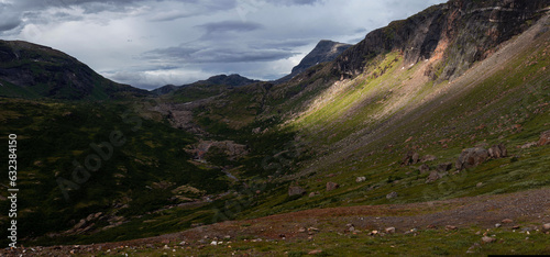 Panorama of sunlit green mountain slopes on the way up to Rabothytta, Okstindan, Hemnes, Helgeland, Northern Norway Green mountains of Nordnorge. Sunny patches on dark mountains. Cloudy dramatic skies photo