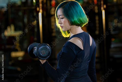 Asian woman is practice weight lifing as beginner on dumbbell for arm and core muscle inside gym with dark background for exercising and workout