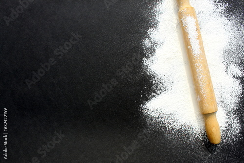 On a black background lies white wheat flour with a wooden rolling pin.