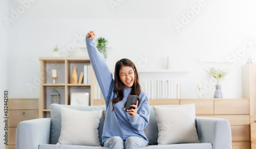 Fotografie Happy young asian woman relax on comfortable couch at home texting messaging on