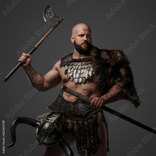 A fierce, muscular, bearded Viking warrior clad in furs and lightweight armor, with a helmet strapped to his waist, wielding two axes against a gray backdrop