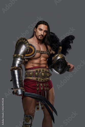 Powerful and attractive gladiator with a stylish beard and luscious locks wears ornate lightweight armor, clutching a gladius and feathered helmet as he stands confidently against a grey backdrop