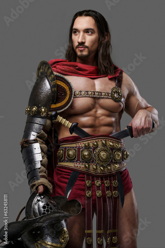 Venerable gladiator in elegant lightweight armor and red cloak poses with helmet, on a grey background