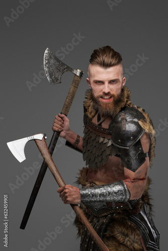 A fierce and bearded Viking warrior dressed in fur and light armor, wielding two axes against a gray background
