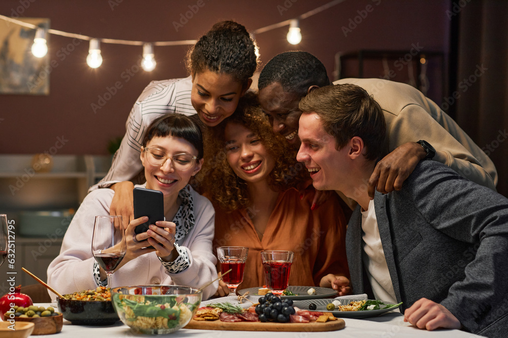 Group of happy young intercultural friends communicating in video chat while looking at screen of smartphone held by smiling brunette girl