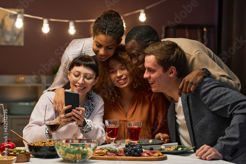 Group of happy young intercultural friends communicating in video chat while looking at screen of smartphone held by smiling brunette girl