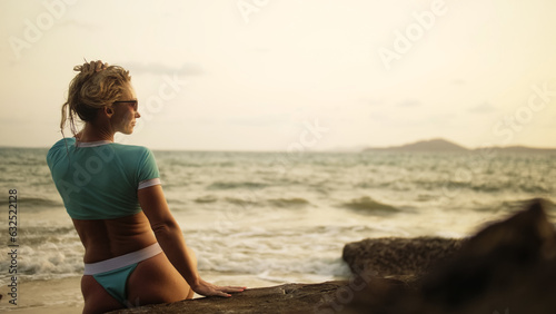 Silhouette sexy woman leaning on a rock reef enjoying and relax warm dark golden sunset. Back view. Woman in blue swimsuit and sunglasses. Concept rest tropical tourism happy summer holidays
