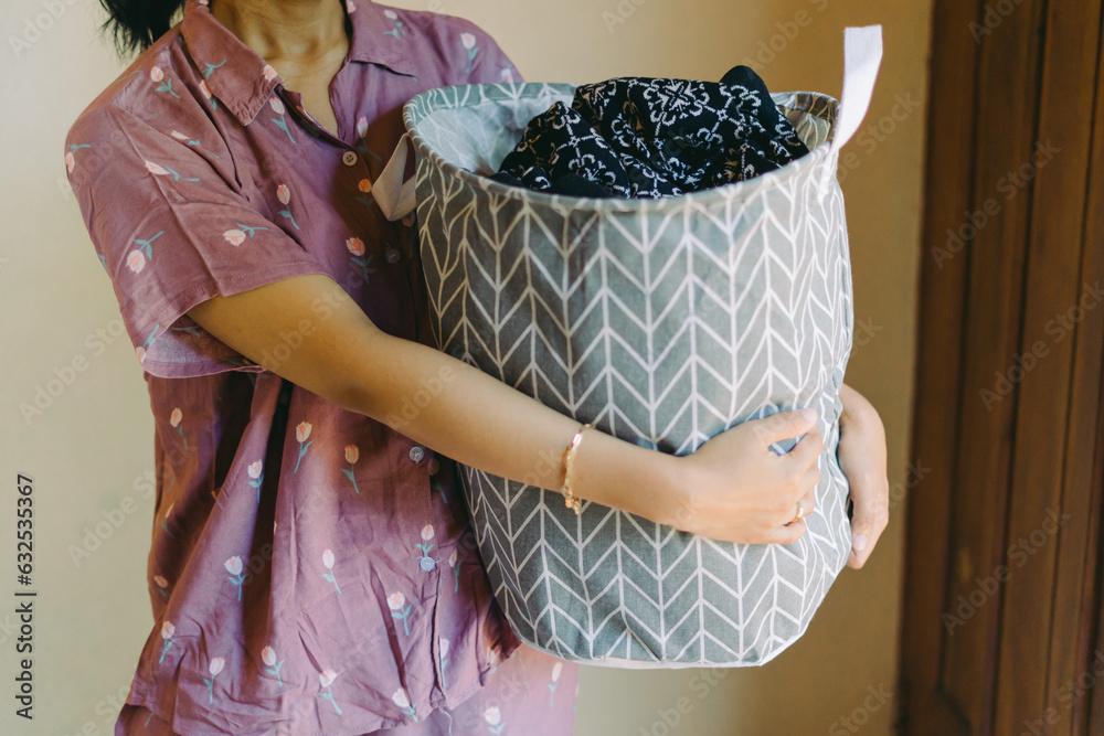 A woman in pajamas carries dirty clothes laundry in a basket to wash