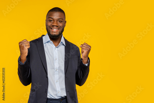 Photo of an african handsome excited businessman dressed with elegant suit, rising clenched fists celebrating victory, isolated over yellow color background at studio.