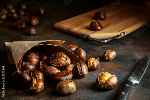 Roasted chestnuts in paper bag on black grunge background. Dark low key photo. Traditional food concept. Selective focus, blurred background.