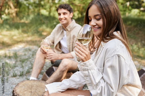 Smiling young woman holding glass of wine and sitting near blurred boyfriend outdoors