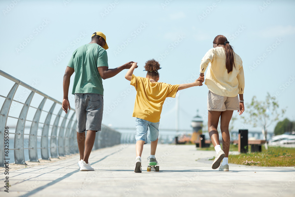 Rear view of little girl learning to skateboarding with her parents outdoors Rear view of little girl learning to skateboarding with her parents outdoors