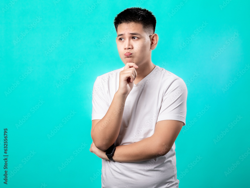 A portrait of an Asian man wearing a white t-shirt, posing as if thinking with his hand on his chin, isolated with a turquoise background.