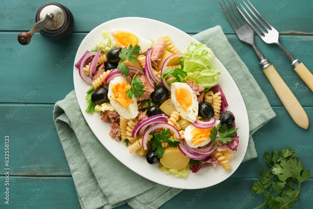Tuna salad with pasta, eggs, potatoes, olives, red onions and sauce in white plate on old turquoise wooden rustic table background. Nicoise salad. French cuisine. Top view. Flat lay. Tuna salad with pasta, eggs, potatoes, olives, red onions and sauce in white plate on old turquoise wooden rustic table background. Nicoise salad. French cuisine. Top view. Flat lay.