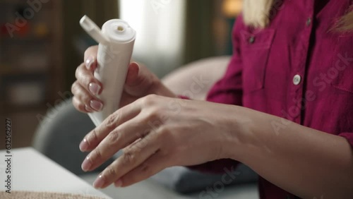 Blonde woman sitting on the sofa, taking a meds tube and applying an ointment to her wrist, close up shot photo