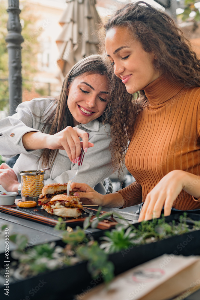 The girl is tasting her friend's burger which looks delicious and yummy.