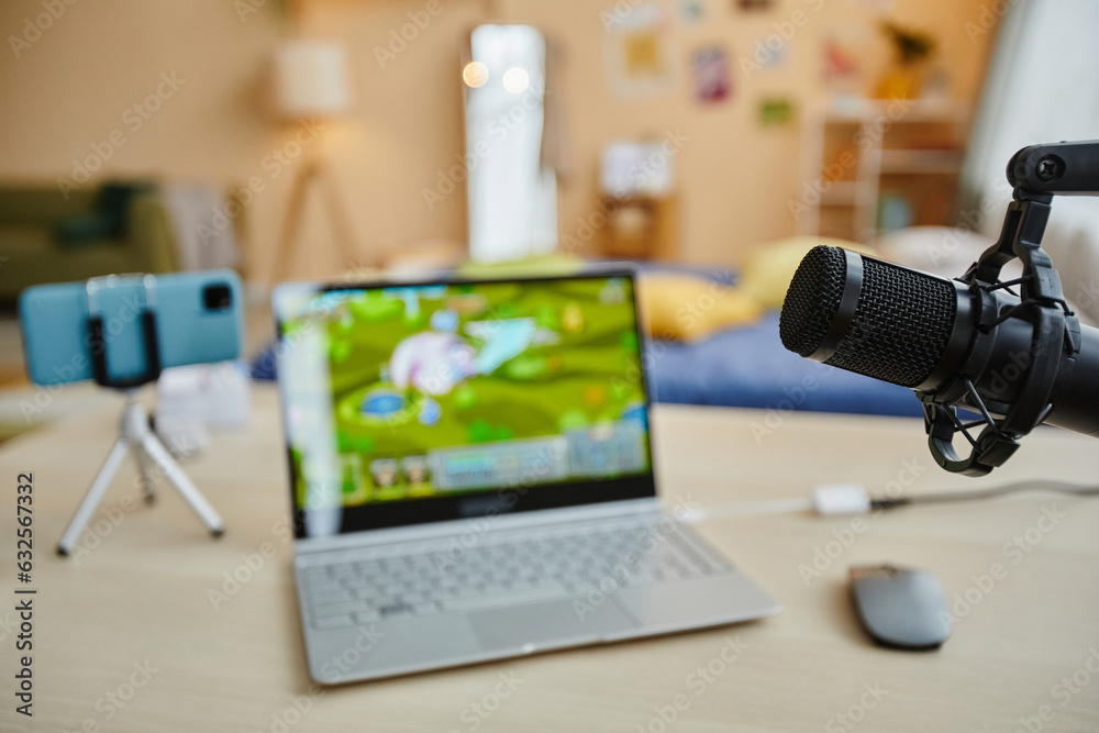 Focus on microphone hanging in front of camera against desk with video game on screen of laptop, smartphone on tripod and desktop mouse Focus on microphone hanging in front of camera against desk with video game on screen of laptop, smartphone on tripod and desktop mouse