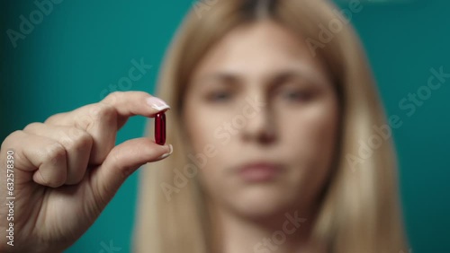 Close up shot of blonde woman model holding a red pill in her fingers, showing it to the camera. Slow motion photo