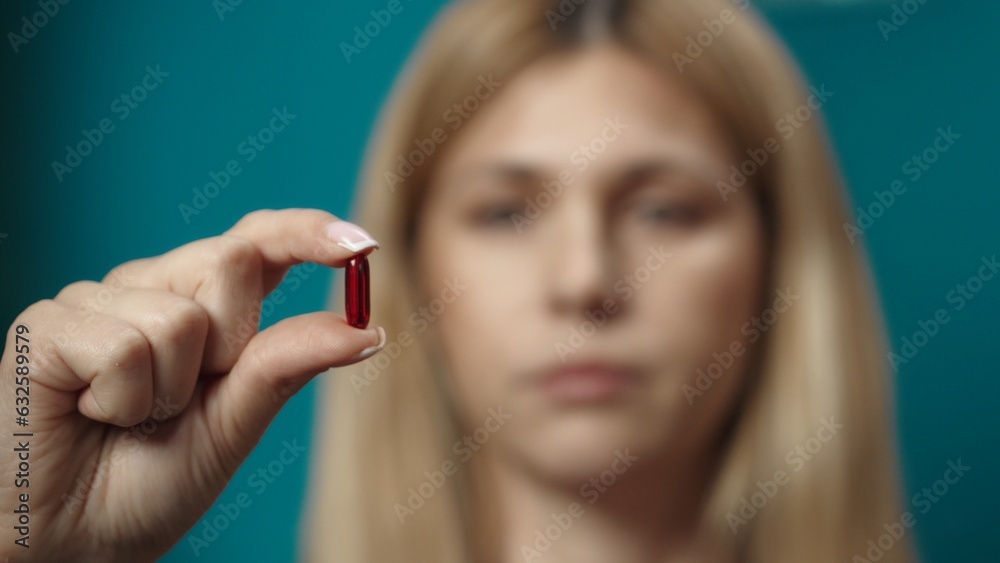 Close up shot of blonde woman model holding a red pill in her fingers, showing it to the camera Close up shot of blonde woman model holding a red pill in her fingers, showing it to the camera