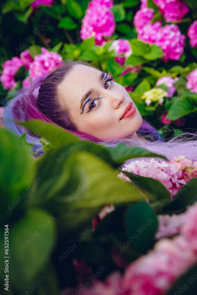 Young woman is surrounded by pink and violet hydrangea flowers. Beautiful model with a long colorful violet hair lying down on blooming hydrangeas flowers in garden Young woman is surrounded by pink and violet hydrangea flowers. Beautiful model with a long colorful violet hair lying down on blooming hydrangeas flowers in garden