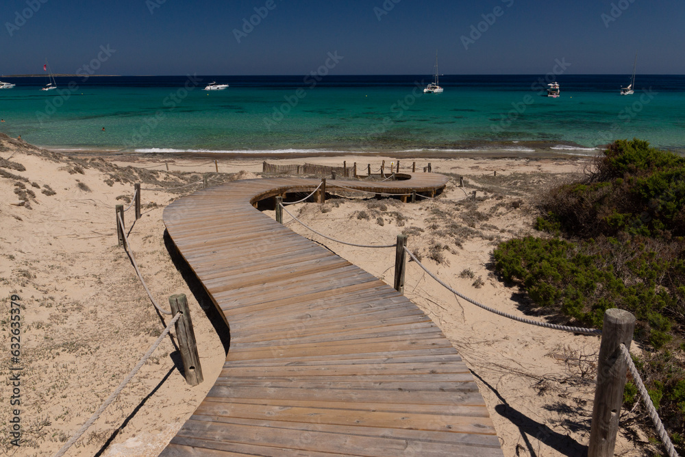 Wooden path to the sandy beach on the island of Formentera and view of the emerald water of the Mediterranean Sea and yachts