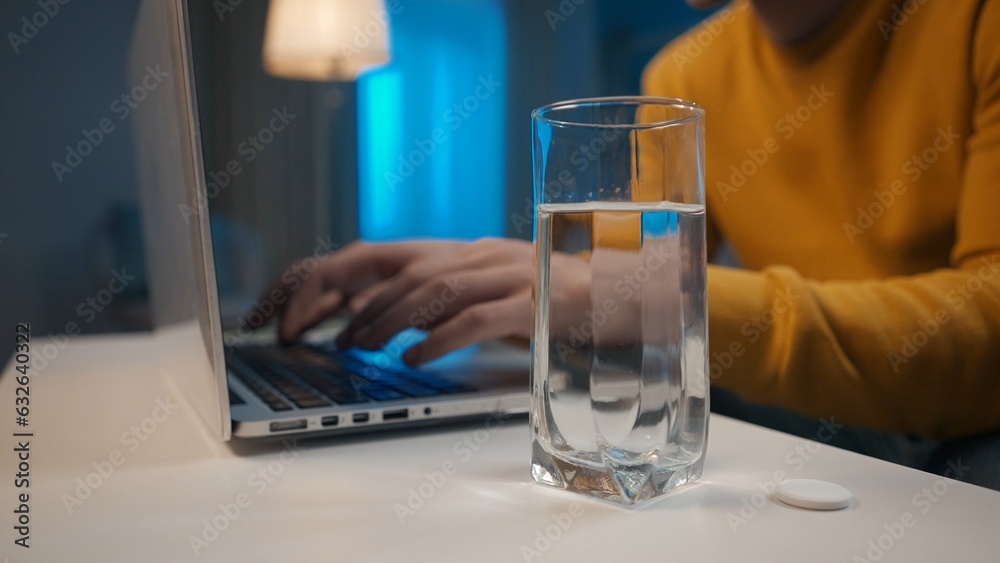 Effervescent tablet and a glass of water on the table close up. In the background, a man's hands are typing on a laptop. Cure for a cold, pain or hangover. Home medicine concept. Effervescent tablet and a glass of water on the table close up. In the background, a man's hands are typing on a laptop. Cure for a cold, pain or hangover. Home medicine concept.