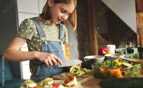 Girl child cuts avicado with knife at kitchen. Pretty female kid with fruits and vegetables preparing salad photo