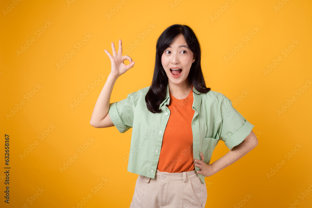 young Asian woman 30s, elegantly clad in orange shirt and green jumper. Her endearing okay hand gesture and gentle smile, set against a yellow background, reveal the beauty of body language. young Asian woman 30s, elegantly clad in orange shirt and green jumper. Her endearing okay hand gesture and gentle smile, set against a yellow background, reveal the beauty of body language.
