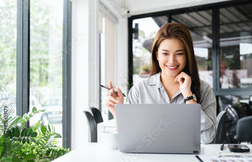 pretty happy young asian entrepreneur woman holding pen working with laptop sitting in the office or cafe.