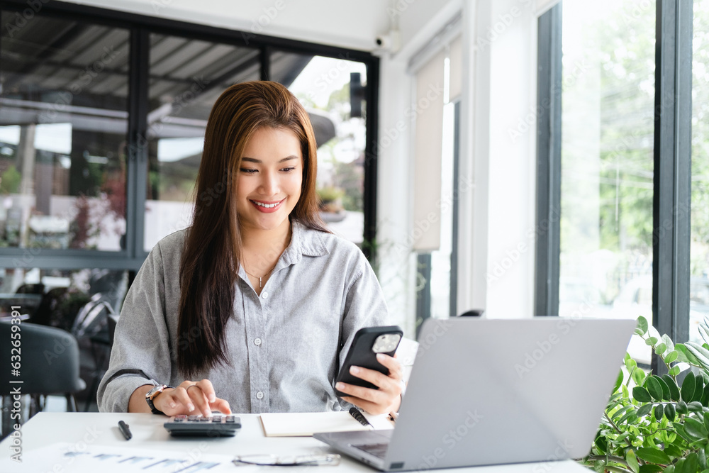 pretty happy young asian entrepreneur woman holding mobile phone working with laptop sitting in the office or co working space cafe.