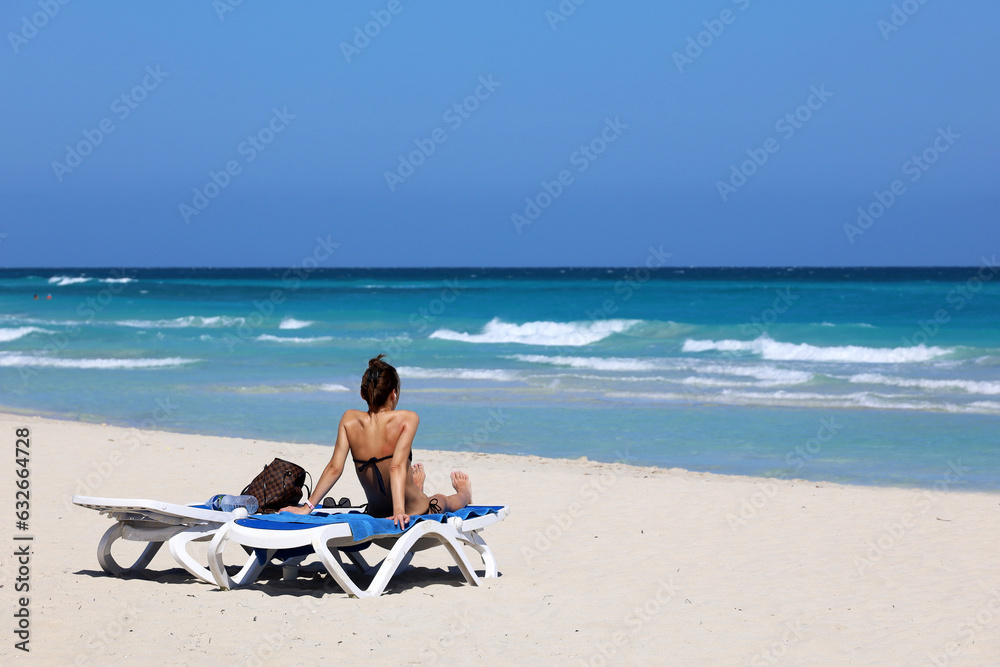 Girl in swimsuit tanning on deck chair on sea waves background. Beach with white sand on ocean coast