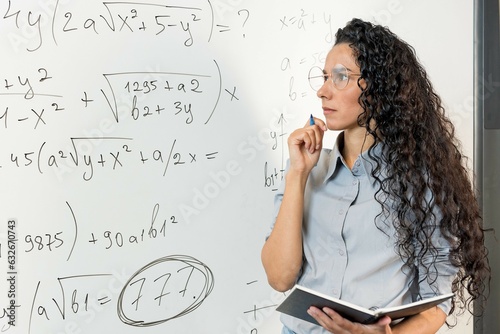 Engaged young Middle Eastern female teacher working on project alone in classroom, looking at whiteboard with math tasks, analyzing notes, thinking over business process schedule, work plan