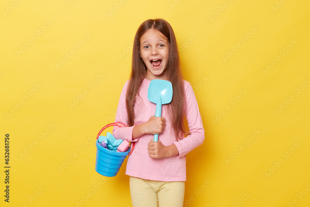 Sunny summer playtime. little cute girl with dark hair holding beach sandbox toys rake shovel and colorful beach bucket. isolated over yellow background screaming with exitement. Sunny summer playtime. little cute girl with dark hair holding beach sandbox toys rake shovel and colorful beach bucket. isolated over yellow background screaming with exitement.