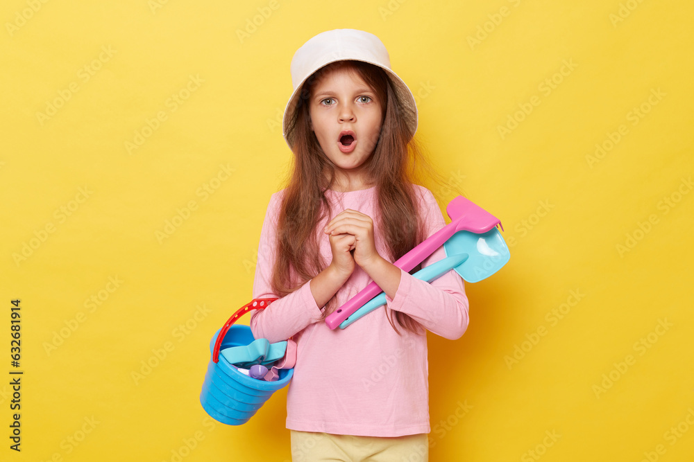 Shocked astonished little cute girl in panama holding beach sandbox toys rake and shovel isolated over yellow background looking at camera with big eyes and open mouth. Shocked astonished little cute girl in panama holding beach sandbox toys rake and shovel isolated over yellow background looking at camera with big eyes and open mouth.