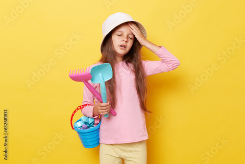 Sad sick unhappy little cute girl in panama holding beach sandbox toys rake and shovel isolated over yellow background suffering heradache spending long hours under sun.