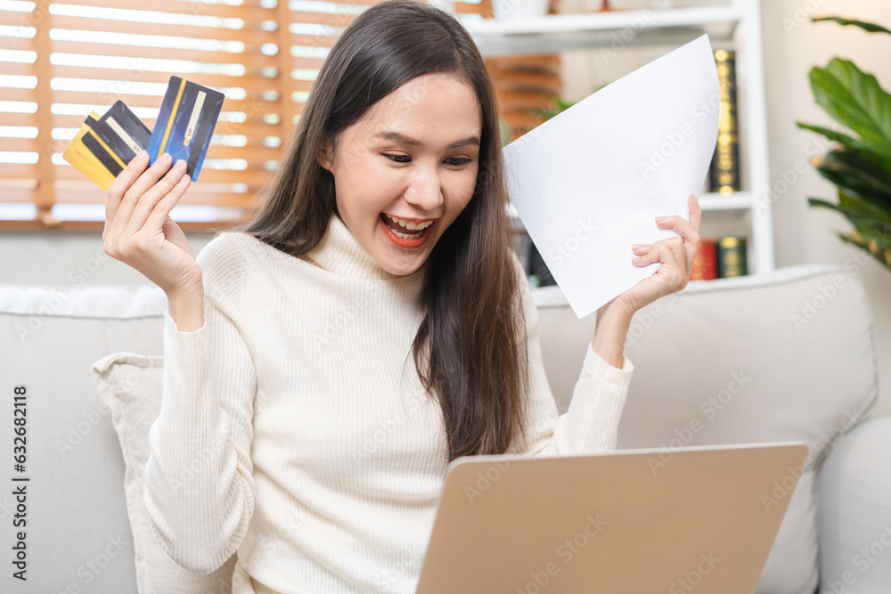 Happy excited, asian young woman, girl holding credit card and paperwork getting, received job promotion, approve tax refund cash back, looking good news at laptop computer, sitting on sofa at home. Happy excited, asian young woman, girl holding credit card and paperwork getting, received job promotion, approve tax refund cash back, looking good news at laptop computer, sitting on sofa at home.