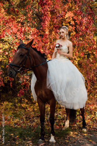 Beautiful bride in white dress stands near a horse in the autumn forest. 