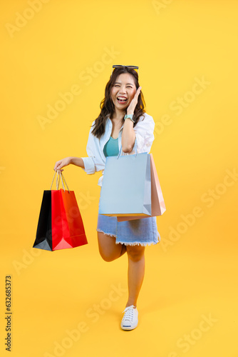 portrait of young Asian woman announcing sale promotion and holding shopping bags enjoy buying gifts in store isolated yellow studio background, happy smile shopper lifestyle spend weekend summer time