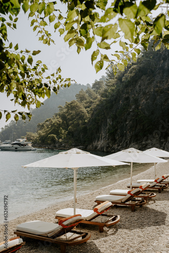 Fototapeta Naklejka Na Ścianę i Meble -  Tranquil beach scene with wooden sun loungers and white parasols lined along a pebbled shore. The turquoise sea, lush green mountains, and anchored yachts create a serene luxury escape in Türkiye