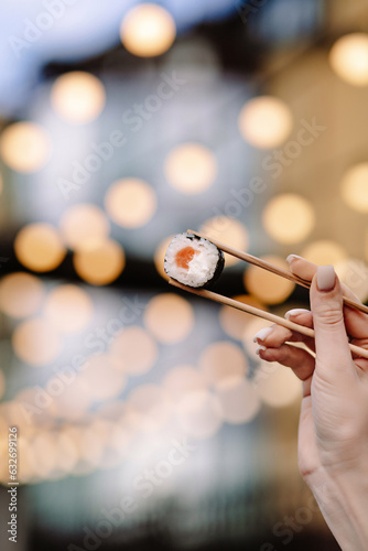 a woman's hand holds a salmon roll on chopsticks with flashlights in the background