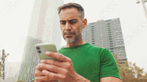 Mature man wearing casual clothes, standing on modern buildings background, texting on smartphone