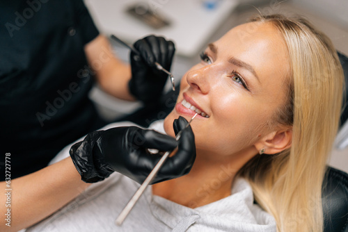 Detail headshot of happy blonde female patient having treatment in dentistry clinic. Close-up hands of unrecognizable dentist in gloves examining patient teeth with dental equipment instrument.