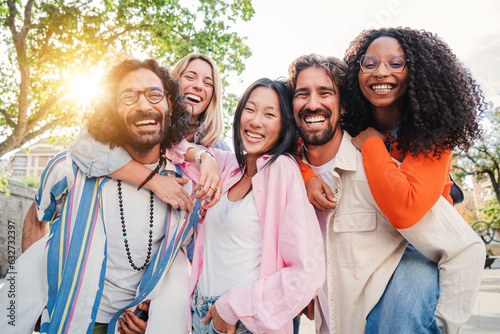 Group of best friends having fun, laughing and smiling taking a selfie portrait outside. Young adult international people celebrate their friendship. Multiracial males giving a piggyback to females