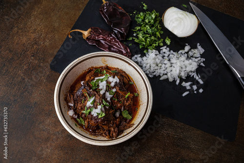 Fototapeta Naklejka Na Ścianę i Meble -  A bowl of slow cooked and braised birria beef meat in a vintage bowl with chopped onions, cilantro, guajillo peppers, and broth