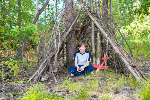 Canvastavla a happy 10 year old boy sits in a makeshift hut made of branches