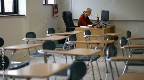 Billede på lærred Happy female teacher sitting at desk in empty school classroom desk grading papers working on assignments